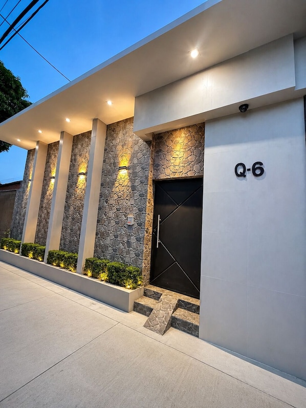 A modern building facade with a stone-textured wall, a black door, and illuminated wall sconces.