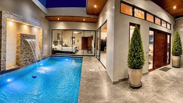 A modern indoor pool area with a waterfall feature, a bedroom with a glass door, and a reception area with a wooden door.