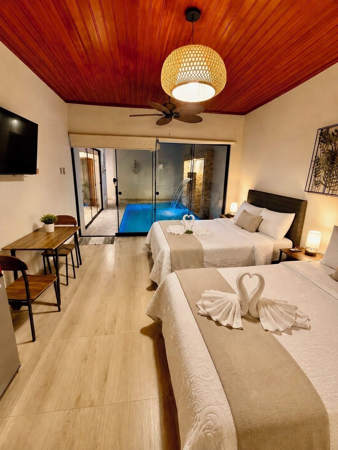 A hotel room with two beds, a dining area, a TV, a ceiling fan, and a view of a swimming pool.