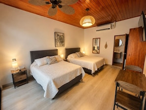 A hotel room with two beds, a wooden ceiling, a ceiling fan, a wall-mounted air conditioner, a bedside table with a lamp, and a mirror.