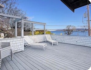 Eine überdachte Terrasse im Freien mit zwei weißen Liegestühlen, einer Pergola und Blick auf ein Gewässer.
