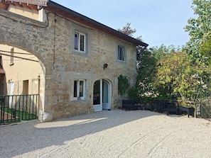 Un bâtiment en pierre avec une allée de gravier, une porte en bois et un petit jardin agrémenté de bancs.