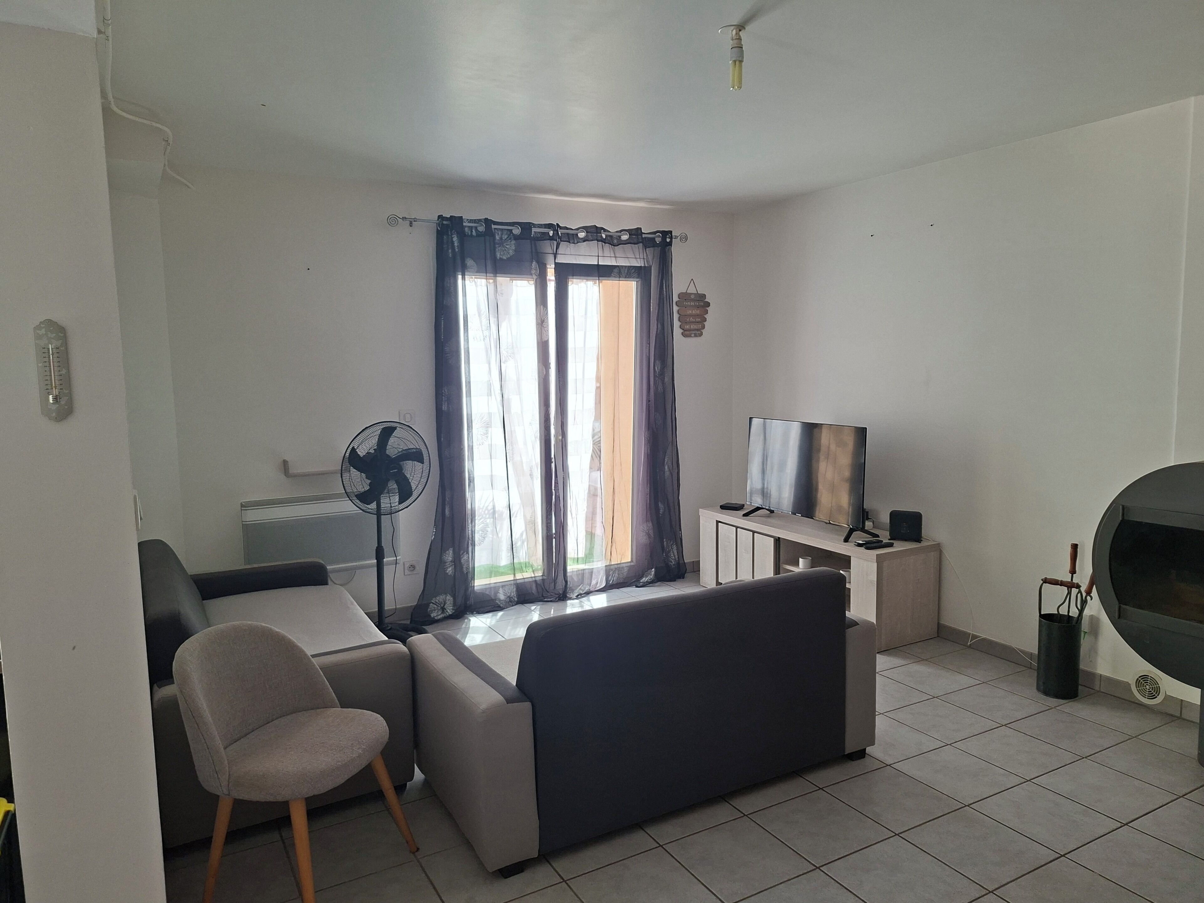 A living room with a sofa, a chair, a TV on a stand, a fan, and a window with curtains.