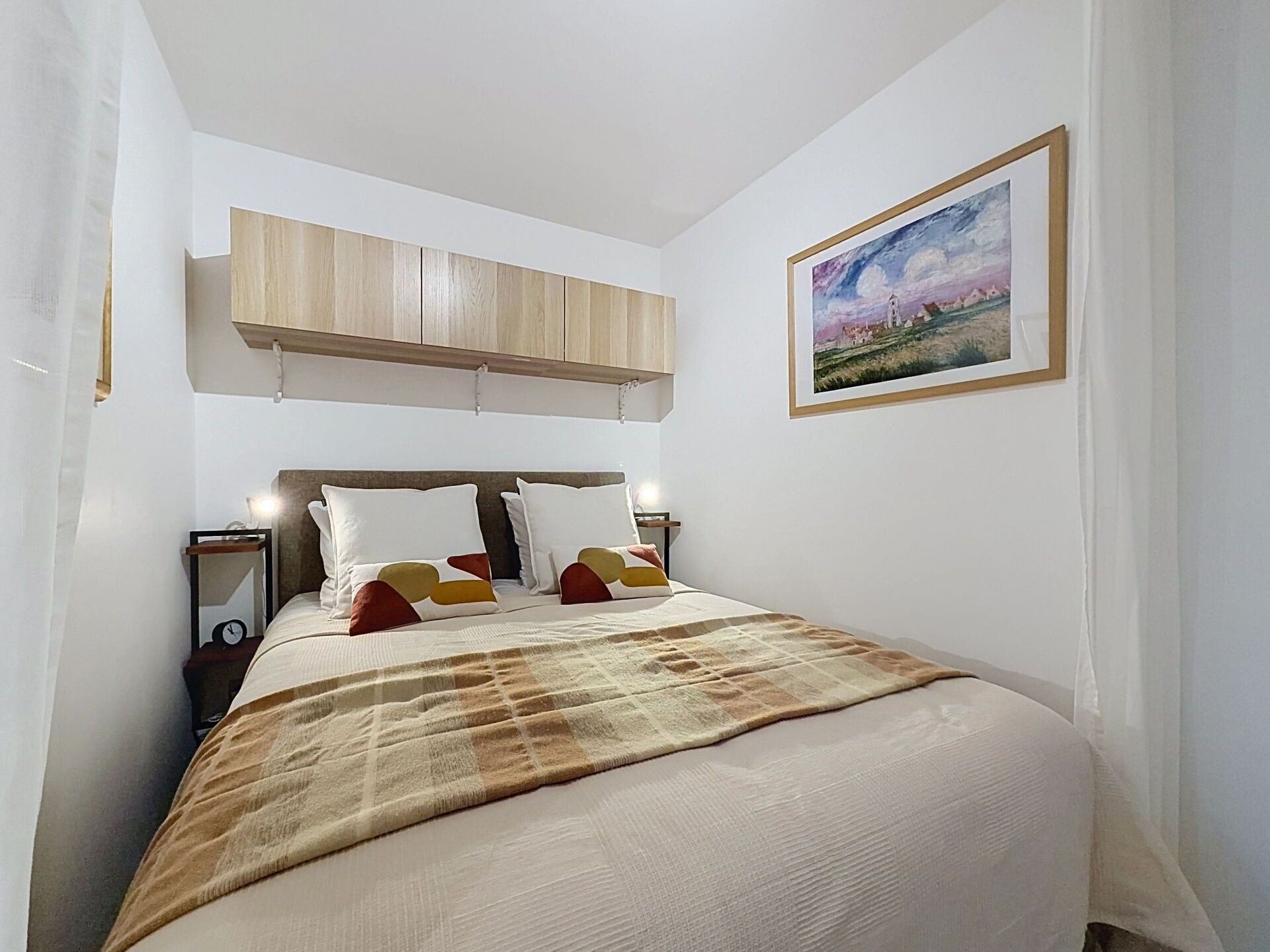 Een slaapkamer met een bed, nachtkastje, schilderij en een houten kast boven het bed.