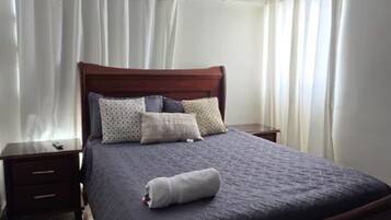 A neatly made bed with a dark-colored bedspread and multiple pillows, a wooden headboard, and a small bedside table with a lamp.