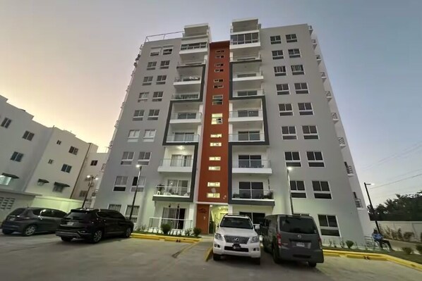 A multi-story residential building with balconies, a parking area with several cars, and a clear sky.