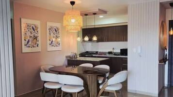 A dining area with a wooden table and white chairs, a kitchen with dark cabinets, and a hanging light fixture.