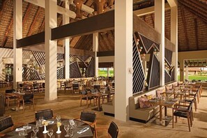 Un vaste espace extérieur aménagé pour les repas, avec de nombreuses tables et chaises, entouré de hauts plafonds aux poutres apparentes et surmonté d’un toit de chaume.
