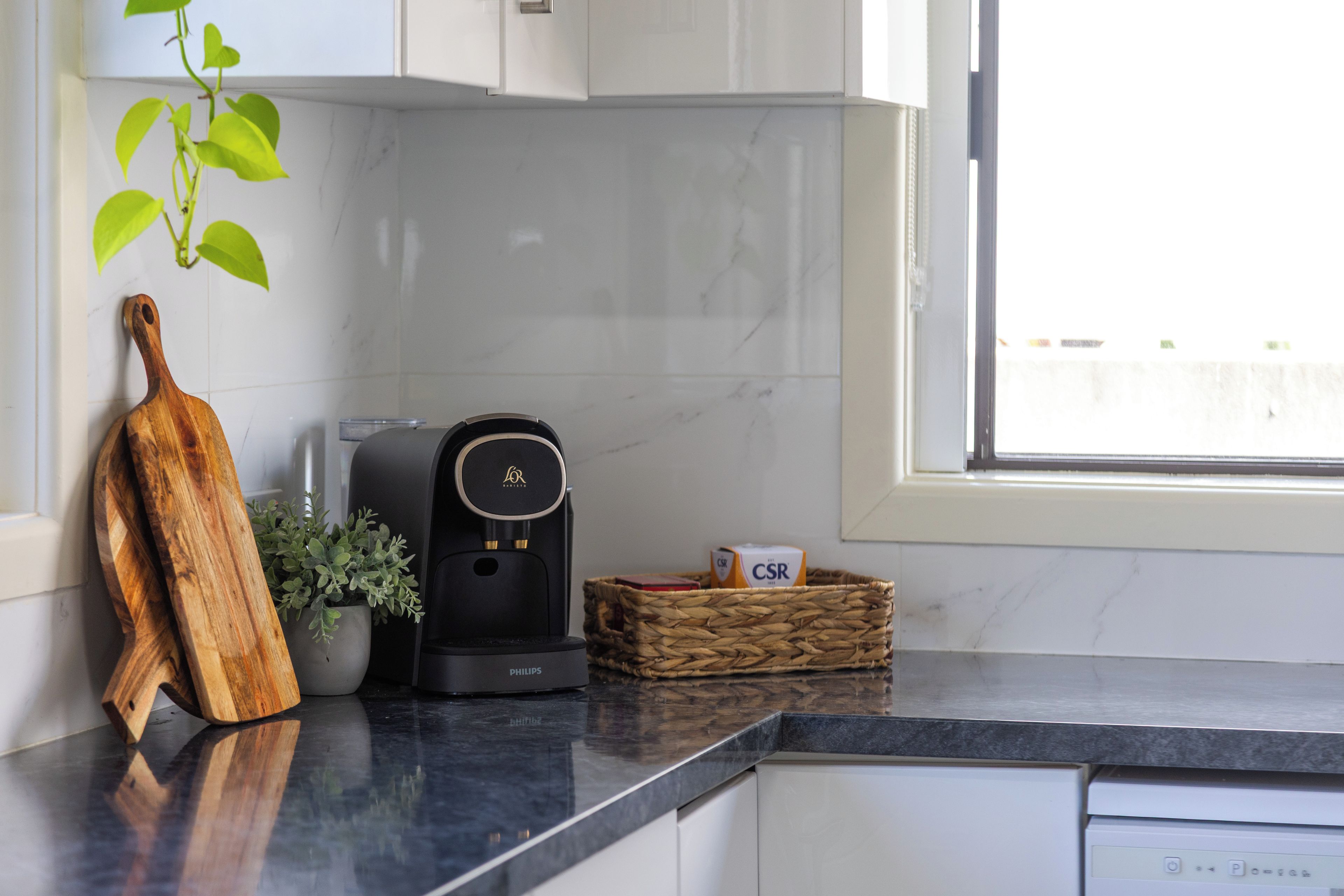 A kitchen with a coffee maker, a wooden cutting board, a potted plant, and a basket with a box labeled 'CSR'.