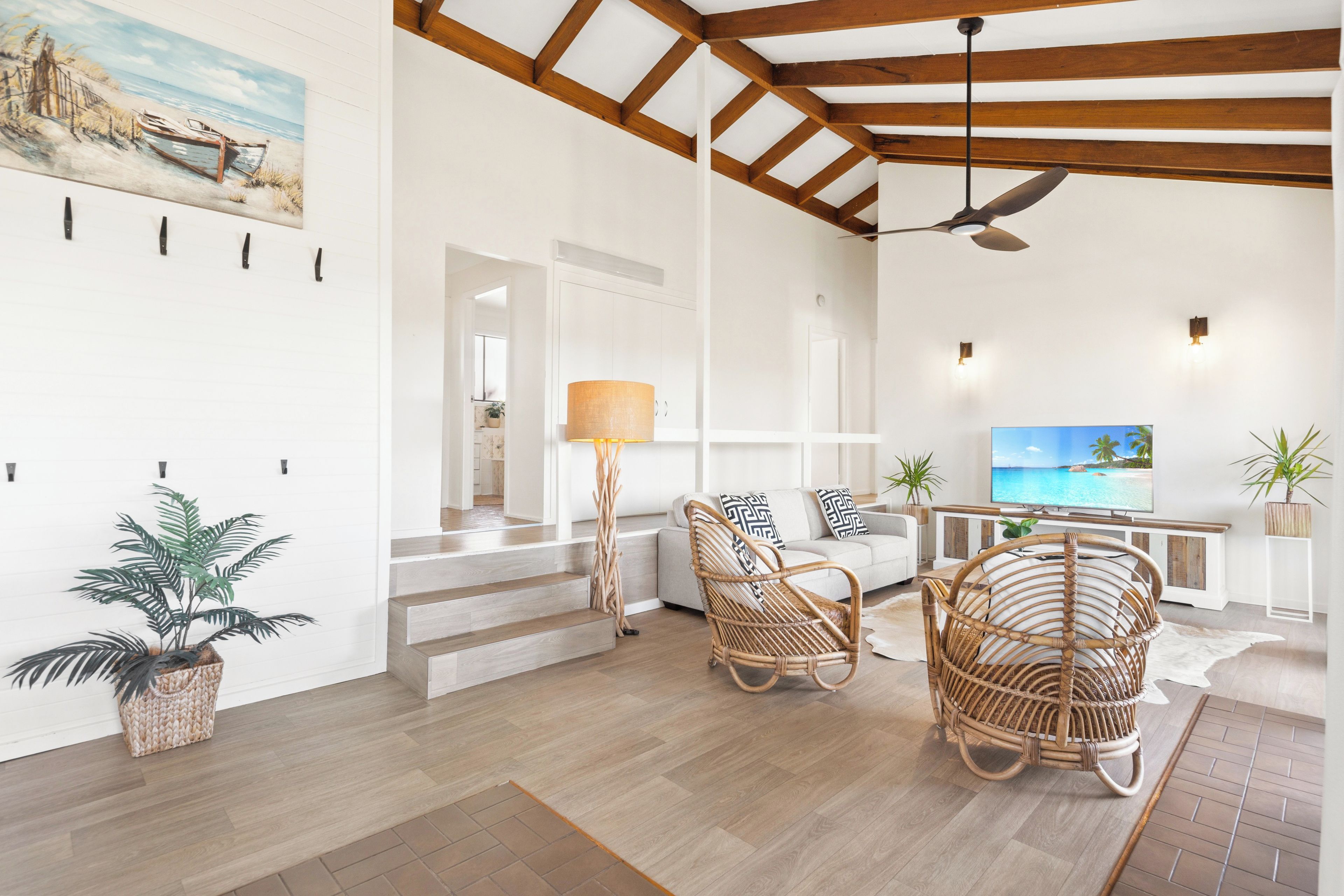 A spacious living area with a high ceiling, wooden beams, a ceiling fan, wicker furniture, a flat-screen TV, and a painting of a beach scene.