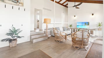 A spacious living area with a high ceiling, wooden beams, a ceiling fan, wicker furniture, a flat-screen TV, and a painting of a beach scene.