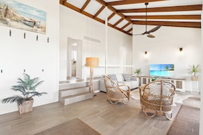 A spacious living area with a high ceiling, wooden beams, a ceiling fan, wicker furniture, a flat-screen TV, and a painting of a beach scene. - Ocean Views - 300m to Beach - 3km to Big Banana (Korora)