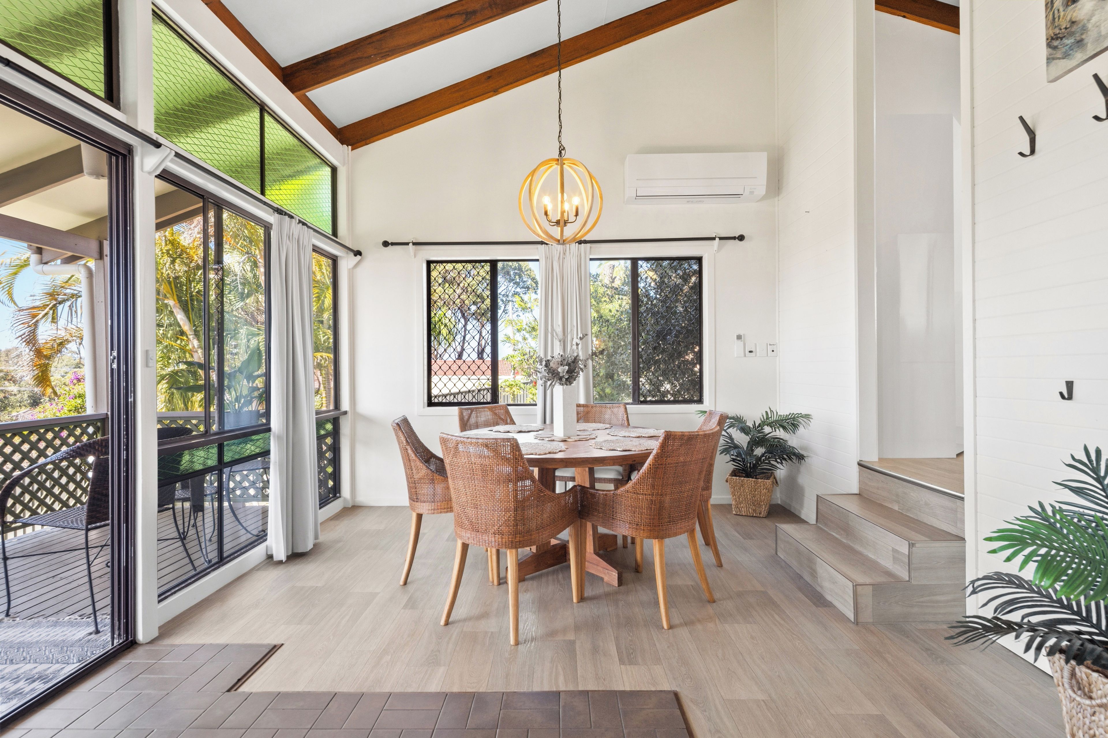 A dining area with a table and chairs, a hanging light fixture, and large windows with a view of greenery.