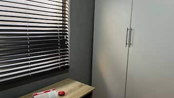 Et værelse med et vindue med vandrette persienner, et træbord med en kalender og en rød genstand, samt en hvid garderobe med to håndtag.