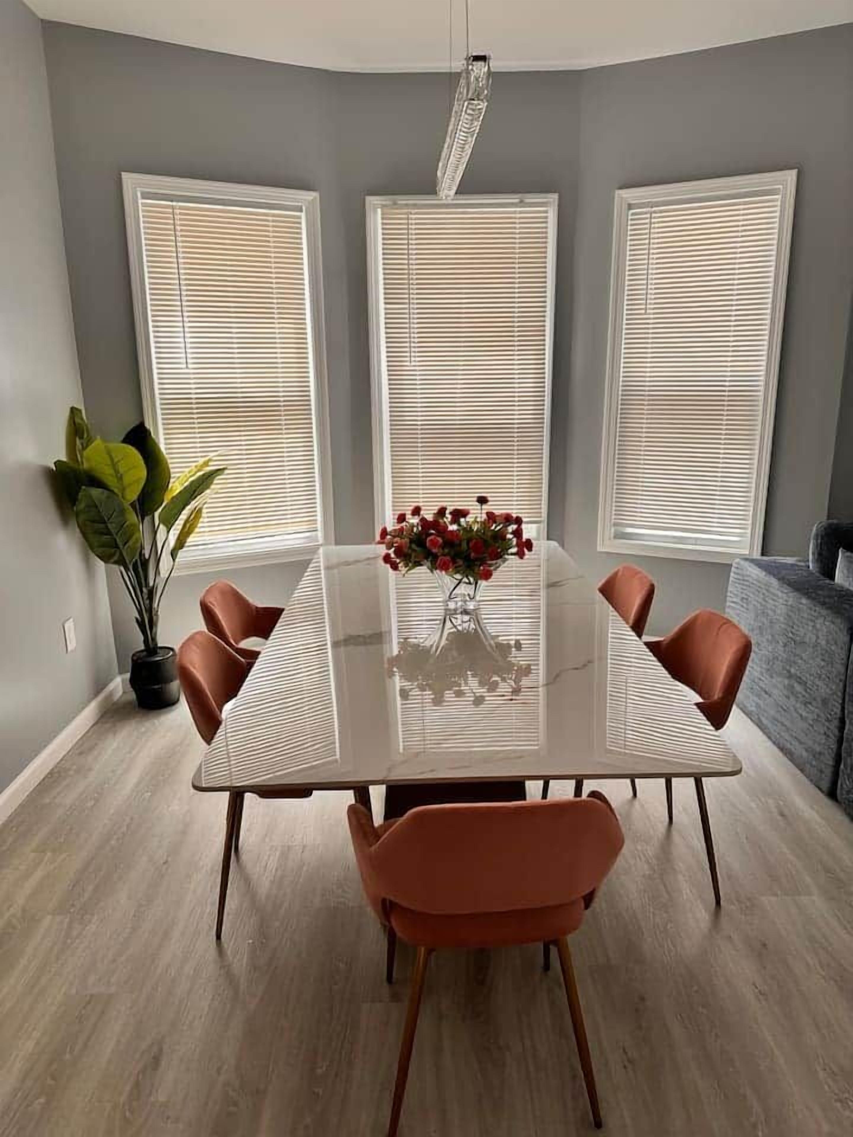 Un comedor con una mesa de vidrio, seis sillas, un jarrón con flores y tres ventanas con persianas.