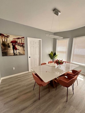 A dining area with a white table, six chairs, a chandelier, a door, and a window with blinds. - Brand New Stunning Home 3 min to Niagara Falls (Niagara Falls)