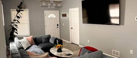 A modern living room with a sectional sofa, a coffee table, and a flat-screen TV mounted on the wall.