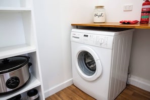 A compact laundry area with a washing machine, a shelf with a jar, and a red fire extinguisher.