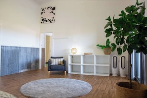 A room with a wooden floor, a grey rug, a blue armchair, a white shelving unit, a wall-mounted lamp, and a large green plant.