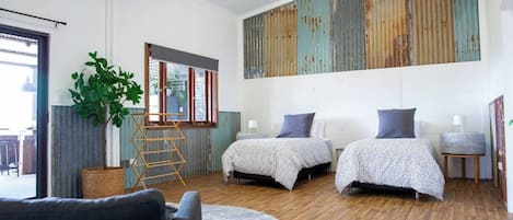 Un dormitorio moderno con dos camas, un ventanal grande, una planta y suelo de madera.