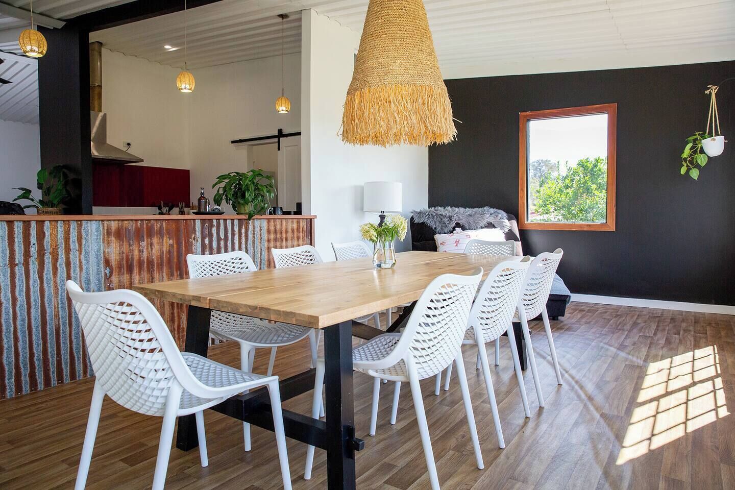 A modern dining area with a wooden table and white chairs, a hanging woven light fixture, and a view of greenery through a window.