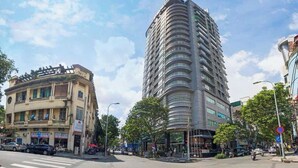 A modern hotel with a curved facade, a ground-level entrance, and a pedestrian area with trees. - IMPERIAL JADE SUITE at THE ONE SAIGON (Ho Chi Minh City)
