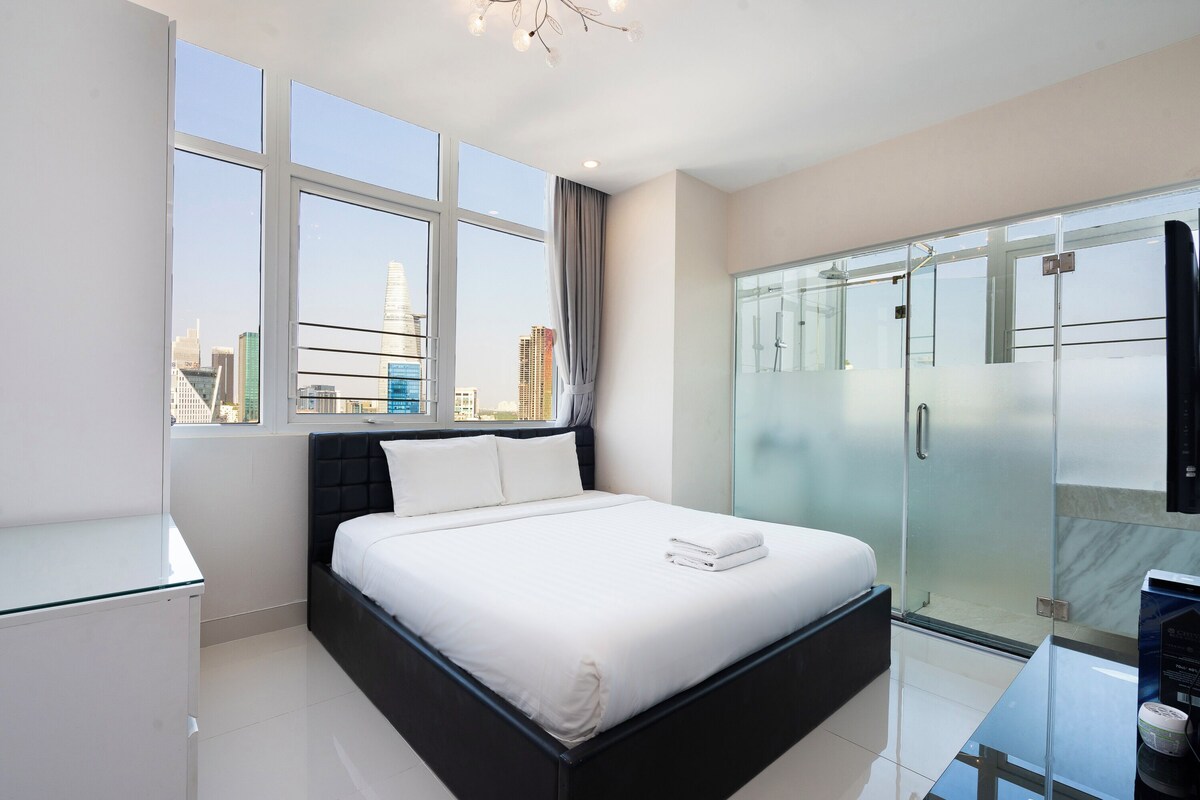 A modern hotel room with a large bed, a glass shower enclosure, and a city view.