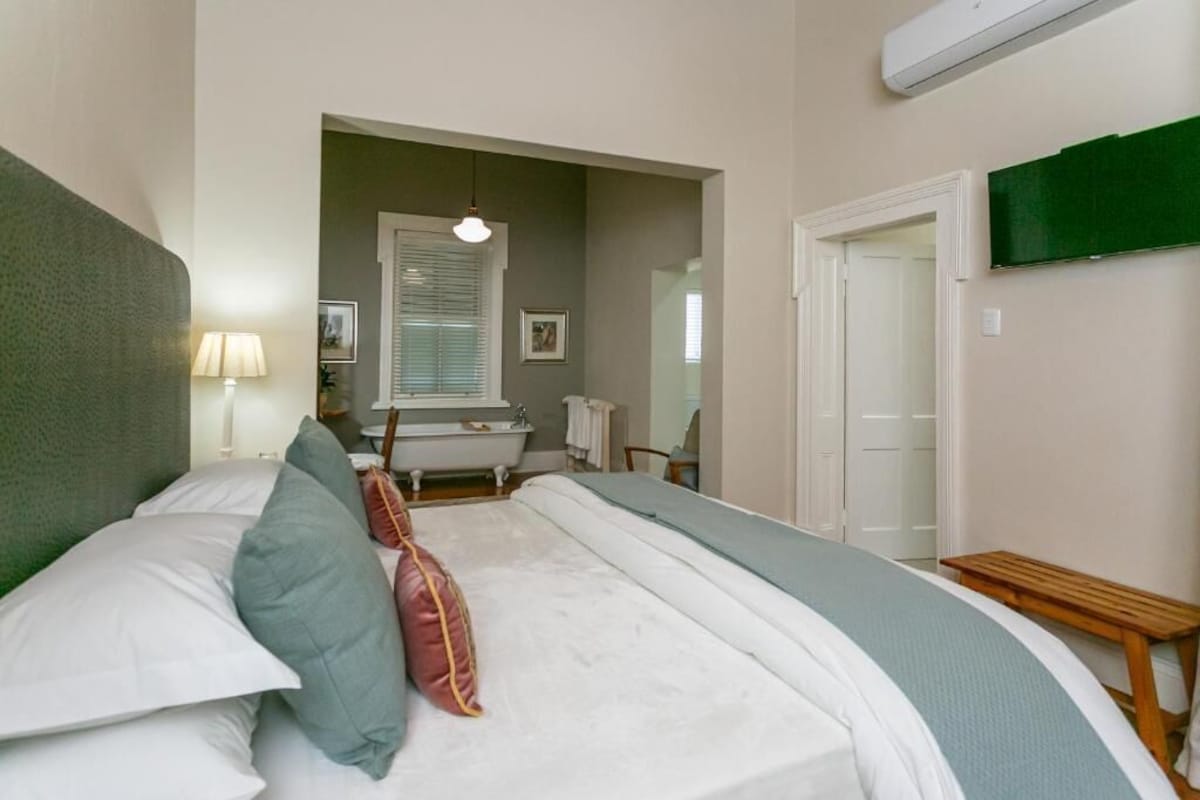 A bedroom with a bed, a bedside lamp, a window with blinds, a mirror, a green wall, and a door.