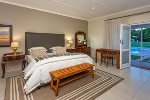 A bedroom with a bed, bedside tables, a bench, a desk, and a view of a swimming pool. - Ostrich Baron's Palace (Oudtshoorn)