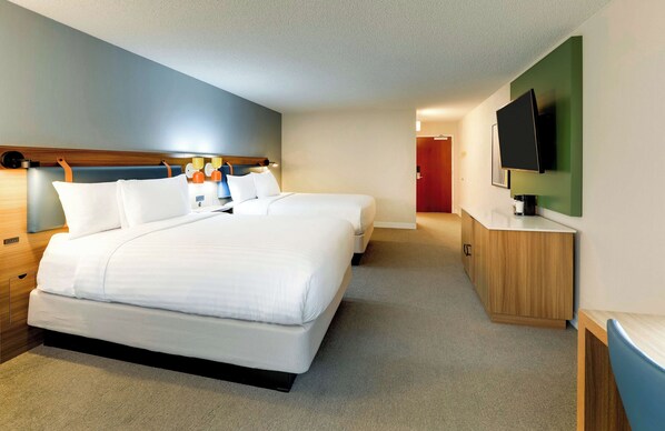 Ein Hotelzimmer mit zwei Betten, einem an der Wand befestigten Fernseher, einem Holzschrank und einer Tür zu einem weiteren Zimmer.