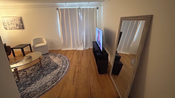 A living room with a flat-screen TV, a white armchair, a glass coffee table, a patterned rug, and a large mirror.