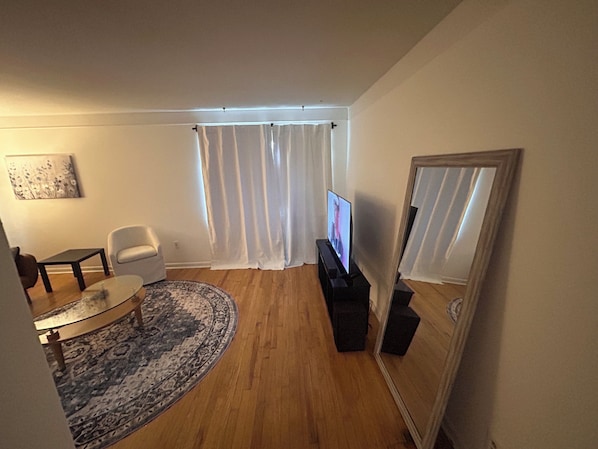 A living room with a flat-screen TV, a white armchair, a glass coffee table, a patterned rug, and a large mirror.