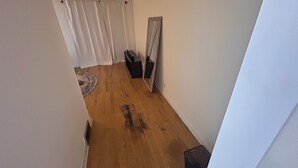 A narrow hallway with wooden flooring, white walls, a black trash bin, a small black object on the floor, and a white curtain at the end.