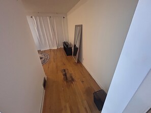 A narrow hallway with wooden flooring, white walls, a black trash bin, a small black object on the floor, and a white curtain at the end.