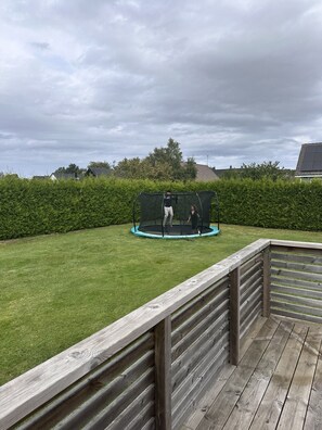 Une terrasse en bois avec un trampoline et un filet de protection, le tout situé dans une cour gazonnée.