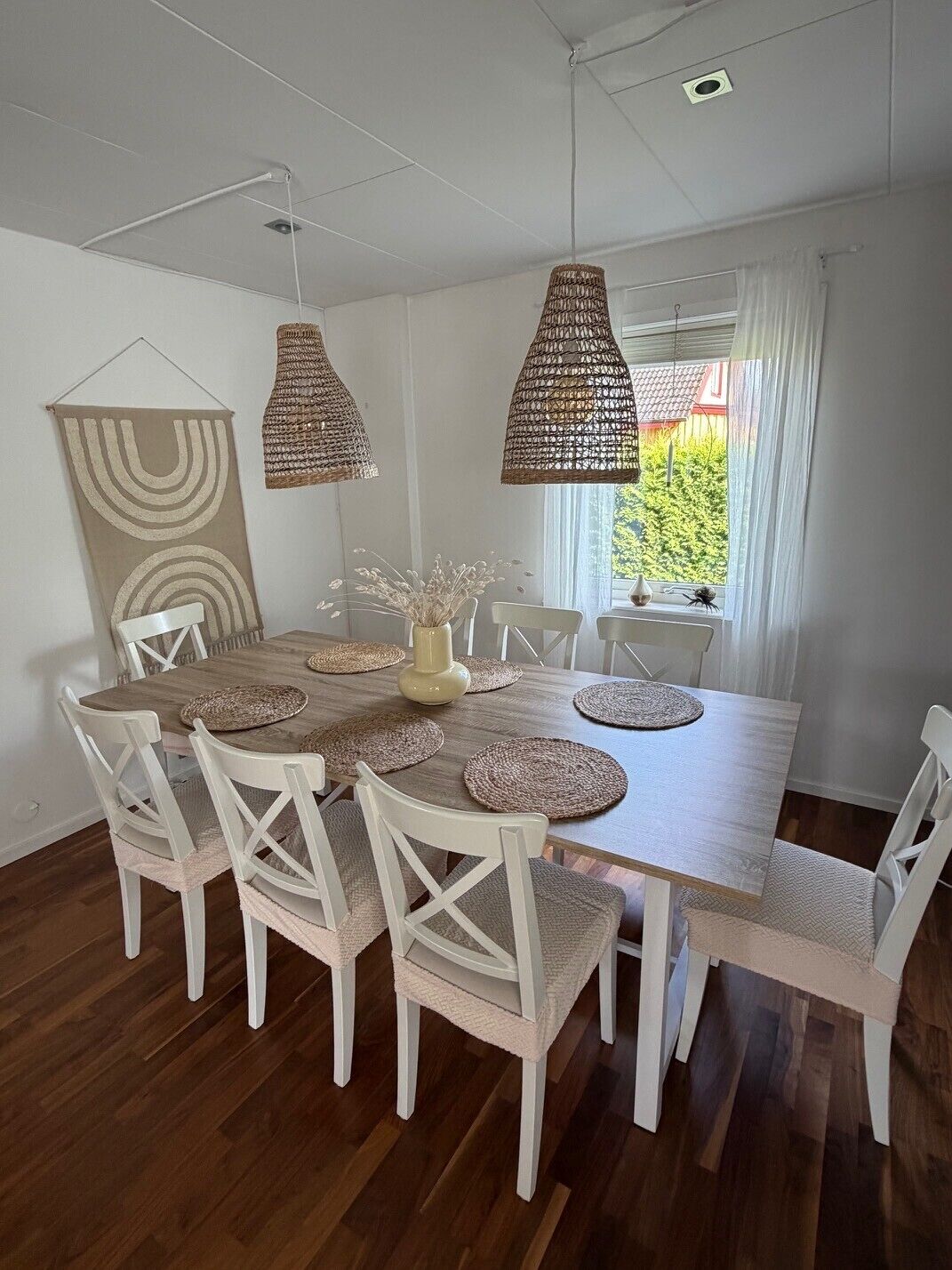 A dining area with a wooden table, white chairs, woven pendant lights, and a view of greenery outside.