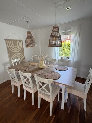 A dining area with a wooden table, white chairs, woven pendant lights, and a view of greenery outside.