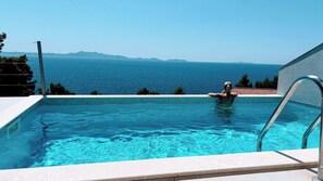 A swimming pool with clear blue water, a metal ladder, and a view of the sea and distant mountains.