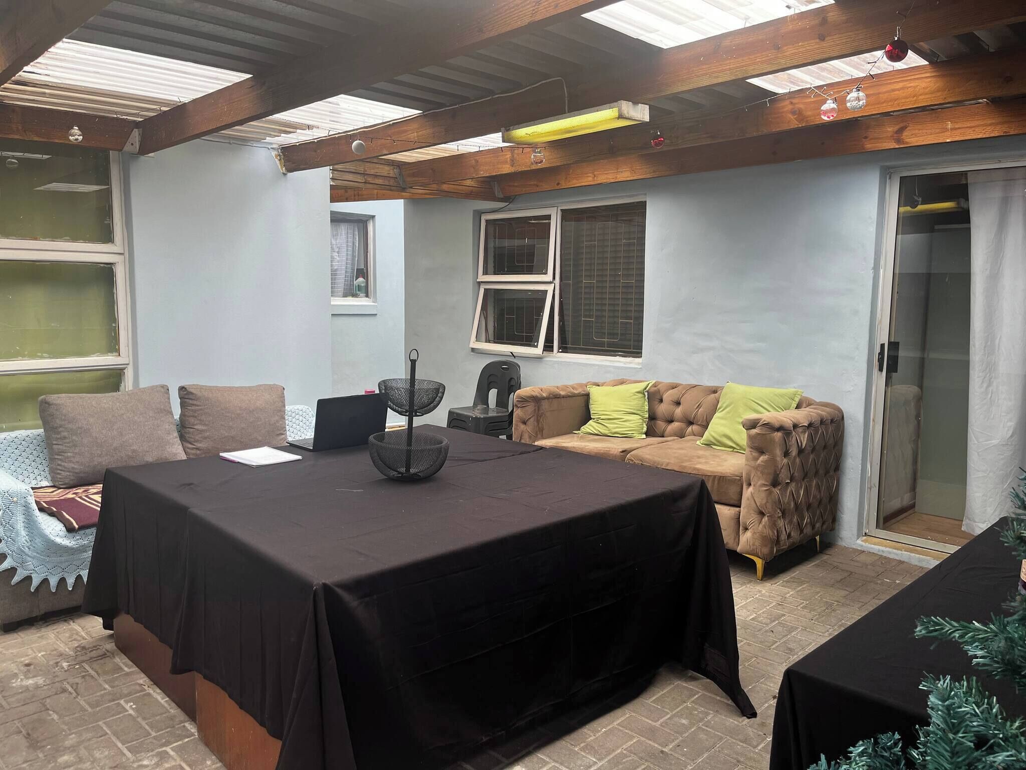 A room with a large table covered with a black tablecloth, a brown sofa with green cushions, and a black chair. There are windows with metal bars and a door leading to a bathroom.