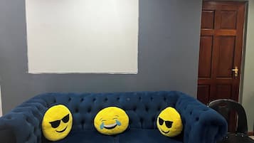 A blue sofa with three yellow smiley face cushions against a grey wall, a blank whiteboard above, and a wooden door with an emergency exit sign.