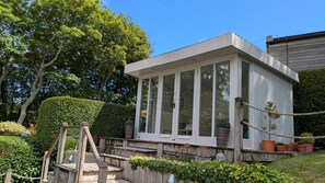 Een oranjerie met een houten terras, omringd door weelderige begroeiing en een helderblauwe lucht.