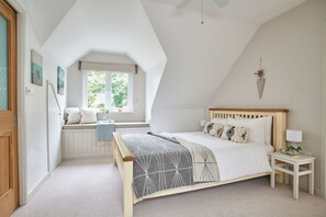 Ein Schlafzimmer mit geneigter Decke, einem Bett mit Holz-Kopfteil, einem Fenster mit Blick auf Grünflächen und einem kleinen Nachttisch mit Lampe.