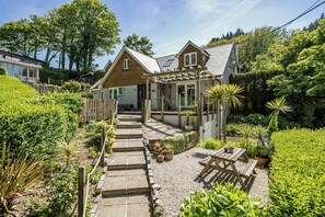 Een huis met een stenen pad dat leidt naar een houten veranda, omringd door weelderig groen en diverse planten.