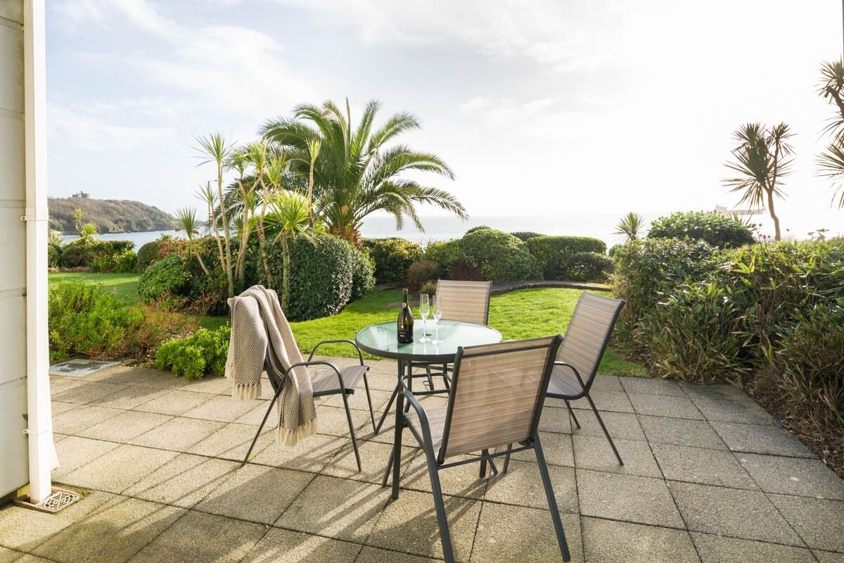 An outdoor patio with a round table and chairs, surrounded by lush greenery and palm trees, overlooking a body of water.