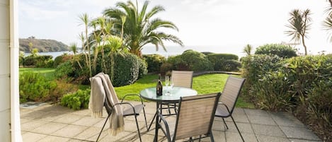 Un patio al aire libre con una mesa redonda y sillas, rodeado de abundante vegetación y palmeras, con vista a un cuerpo de agua.