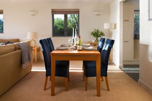 A dining area with a table set for two, two chairs, a window with a view of greenery, a lamp, and a sofa in the background. - Castle Bay - sleeps 4 guests  in 2 bedrooms (Falmouth)