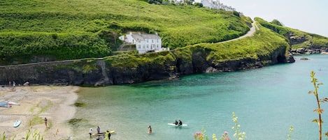 A coastal landscape with a white building on a grassy hill, a clear blue sea, and a rocky shoreline.