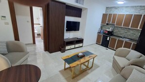 A living room with a TV, sofa, coffee table, and a kitchenette with cabinets and a refrigerator.