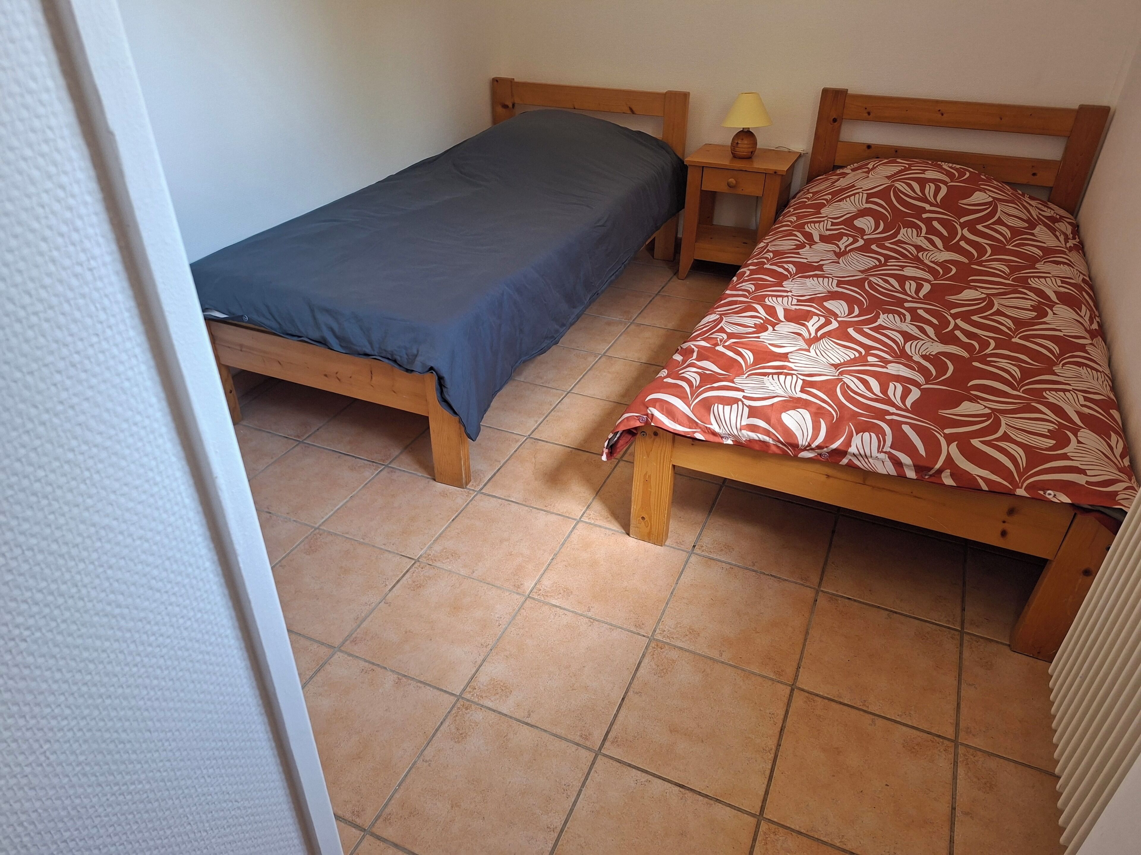 Une petite pièce avec deux lits en bois, l’un recouvert d’une couverture bleue et l’autre d’une couverture rouge à motifs. Une petite table de chevet en bois avec une lampe se trouve entre les deux lits.