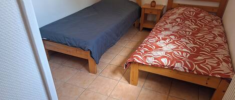 Une petite pièce avec deux lits en bois, l’un recouvert d’une couverture bleue et l’autre d’une couverture rouge à motifs. Une petite table de chevet en bois avec une lampe se trouve entre les deux lits.